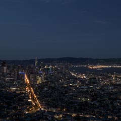 A city skyline at night, illuminated by numerous lights from buildings and vehicles along a busy street.