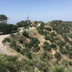 A small hill rises up into scrubby terrain, with a utility tower mounted on top behind a low concrete building with a flat roof, and surrounded by trees.