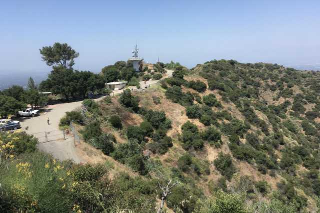 A small hill rises up into scrubby terrain, with a utility tower mounted on top behind a low concrete building with a flat roof, and surrounded by trees.