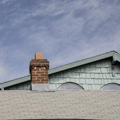 An old red brick chimney rises from a gray shingled roof, with a light green gable and trim visible behind it.