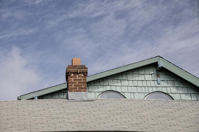 An old red brick chimney rises from a gray shingled roof, with a light green gable and trim visible behind it.