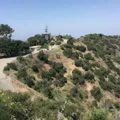A small hill rises up into scrubby terrain, with a utility tower mounted on top behind a low concrete building with a flat roof, and surrounded by trees.