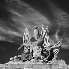 A stone monument featuring a central eagle, cannons, flags, and a shield, set against a dramatic sky.