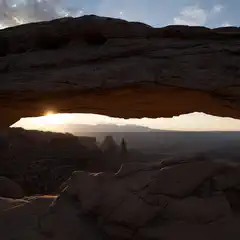 A natural sandstone arch frames the setting sun over a vast desert landscape.