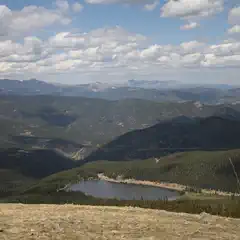 A landscape featuring mountains, forested hillsides, a lake, and a blue sky with clouds.