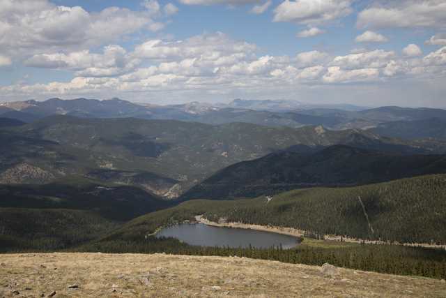 A landscape featuring mountains, forested hillsides, a lake, and a blue sky with clouds.