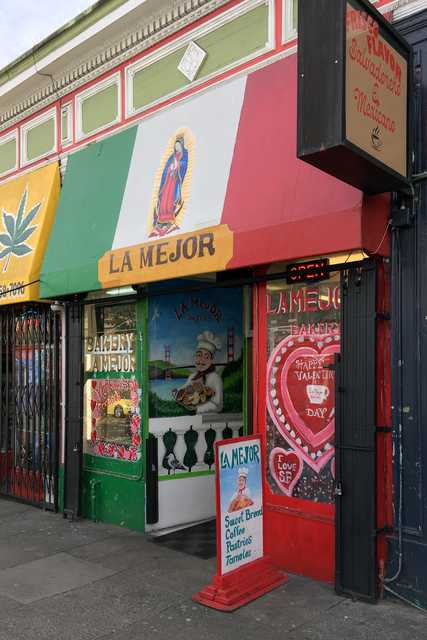 A colorful storefront with a sign that reads "LA MEJOR," featuring an image of Mary and the Mexican flag, along with other decorations and signs.