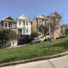 A row of Victorian houses on a sloping street with parked cars and bare trees, under a clear sky.