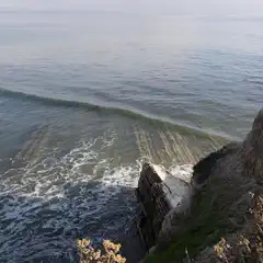 A rocky coastal cliff overlooks a calm ocean with gentle waves breaking near the shore.