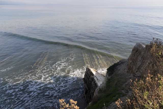 A rocky coastal cliff overlooks a calm ocean with gentle waves breaking near the shore.