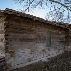 Rustic log cabin with weathered exterior and single window, surrounded by bare trees and hint of water in background.
