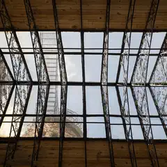 A geometric lattice of metal and glass frames a view of the sky, contrasting with a wooden ceiling and textured stone wall.