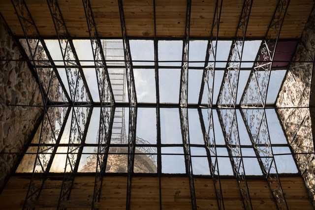 A geometric lattice of metal and glass frames a view of the sky, contrasting with a wooden ceiling and textured stone wall.