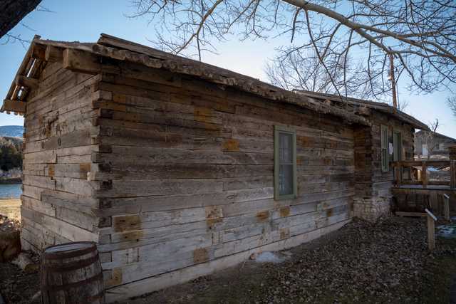 Rustic log cabin with weathered exterior and single window, surrounded by bare trees and hint of water in background.