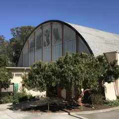 A large, white building with arched windows and a curved roof, topped with a gray shingle roof, surrounded by green leafy trees and bushes on a clear day.
