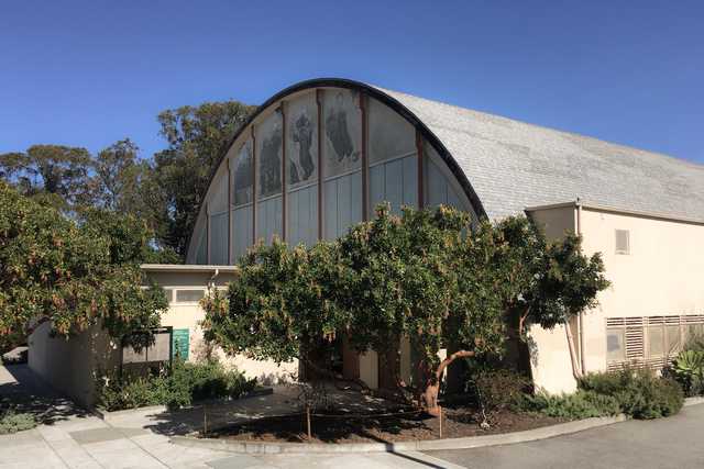 A large, white building with arched windows and a curved roof, topped with a gray shingle roof, surrounded by green leafy trees and bushes on a clear day.