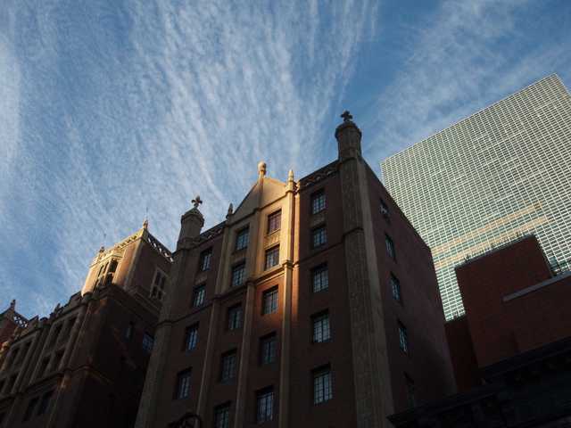 A Gothic-style building with two towers and a central spire rises against a blue sky. A modern glass skyscraper stands in the background.