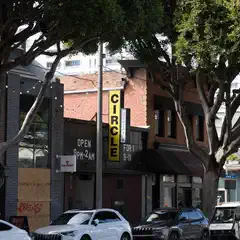 A city street lined with trees and buildings, featuring a bar sign that reads "CIRCLE".