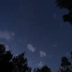 An orange-red streak is visible against a dark blue sky with scattered white clouds, surrounded by silhouetted trees at the bottom and top of the frame.