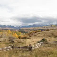 A rustic wooden fence enclosing a dry grass field with trees and mountains in the background under overcast skies.