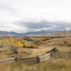A rustic wooden fence enclosing a dry grass field with trees and mountains in the background under overcast skies.