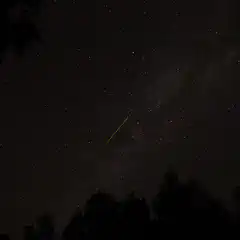 A bright streak of light crosses through a starry sky over a forested area at night.