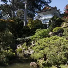 A white Japanese-style pagoda is visible behind a hill covered with green bushes and large rocks.