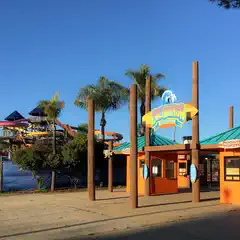 A brightly colored ticket booth stands in front of a large water slide structure featuring multiple spiral slides and a tall, pointed roof.