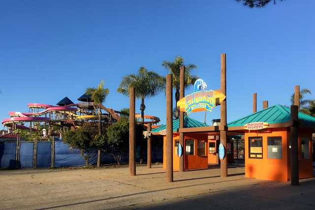 A brightly colored ticket booth stands in front of a large water slide structure featuring multiple spiral slides and a tall, pointed roof.
