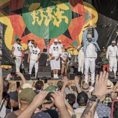 A group of men wearing white clothing stand on stage in front of a large banner with a green circle and yellow figures.