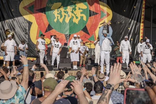 A group of men wearing white clothing stand on stage in front of a large banner with a green circle and yellow figures.