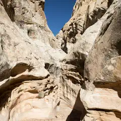 A canyon carved into sandstone rock creates a narrow passage with two towering vertical walls. Steep, rocky cliffs meet in a V-shape against a bright blue sky.