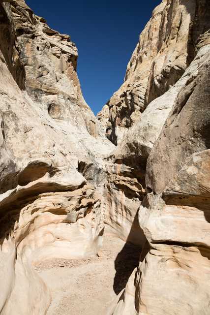 A canyon carved into sandstone rock creates a narrow passage with two towering vertical walls. Steep, rocky cliffs meet in a V-shape against a bright blue sky.