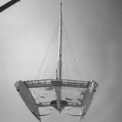 A catamaran is suspended in mid-air by a crane, with a large trailer and people visible below.
