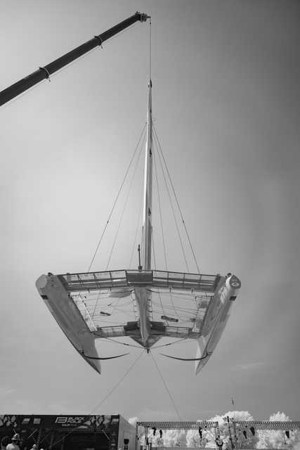 A catamaran is suspended in mid-air by a crane, with a large trailer and people visible below.