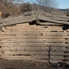An old, rustic log cabin with visible gaps between logs and a weathered appearance, set against a backdrop of dry grass and distant mountains under a clear sky.