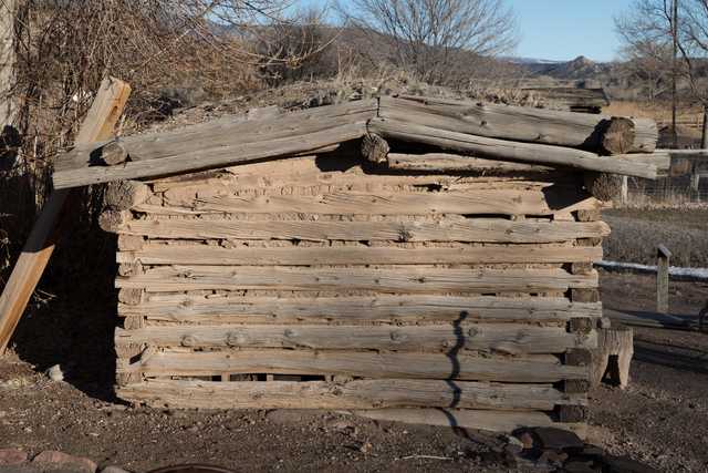 An old, rustic log cabin with visible gaps between logs and a weathered appearance, set against a backdrop of dry grass and distant mountains under a clear sky.