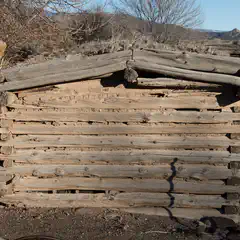 An old, rustic log cabin with visible gaps between logs and a weathered appearance, set against a backdrop of dry grass and distant mountains under a clear sky.