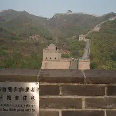 An ancient watchtower stands atop a stone wall, surrounded by lush greenery and distant mountains.