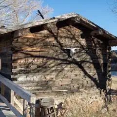 A weathered wooden cabin stands alongside a body of water, with a prominent skull mounted on its side.