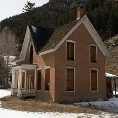 An abandoned, weathered brick house with a sloping roof and missing windows, surrounded by snow-covered ground and bare trees on a mountainous landscape.