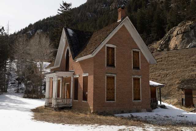 An abandoned, weathered brick house with a sloping roof and missing windows, surrounded by snow-covered ground and bare trees on a mountainous landscape.