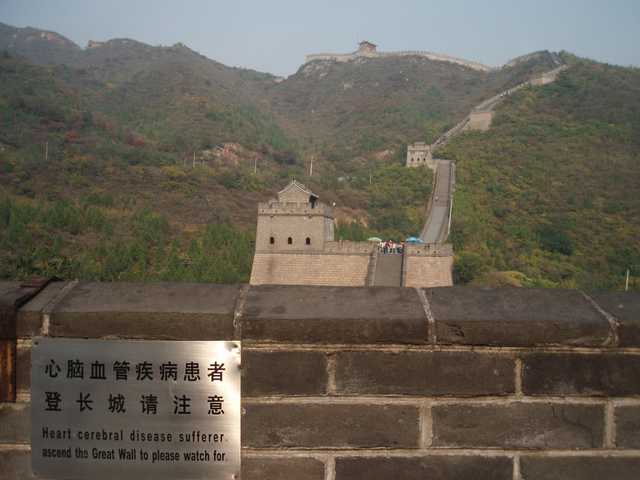 An ancient watchtower stands atop a stone wall, surrounded by lush greenery and distant mountains.