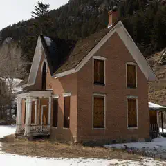 An abandoned, weathered brick house with a sloping roof and missing windows, surrounded by snow-covered ground and bare trees on a mountainous landscape.