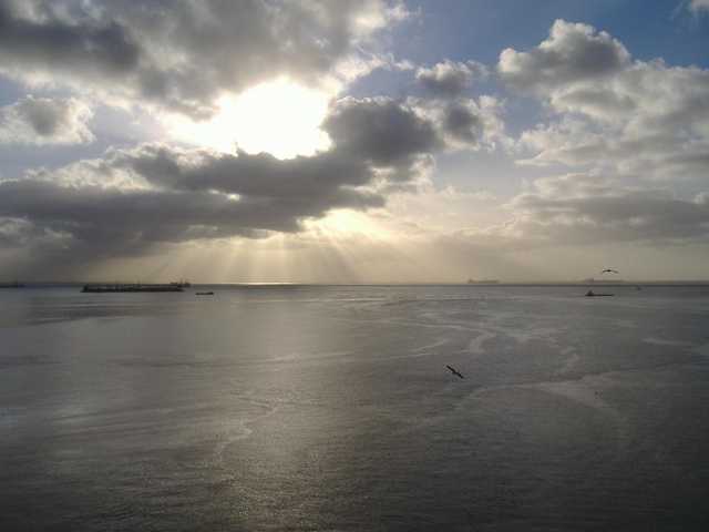Sunlight piercing through clouds over calm water with distant boats and birds.