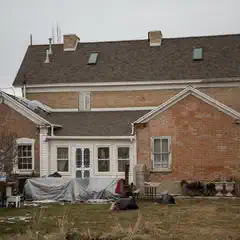 A large, two-story brick house with a shingled roof and multiple windows, surrounded by a yard with sparse grass and scattered debris.