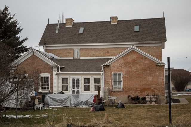 A large, two-story brick house with a shingled roof and multiple windows, surrounded by a yard with sparse grass and scattered debris.