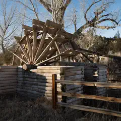 An old wooden waterwheel is situated behind a log fence, with its spokes and paddles visible.