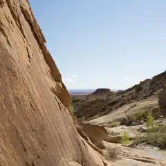 An arid landscape features large sandstone rock formations and a small stream in the foreground.
