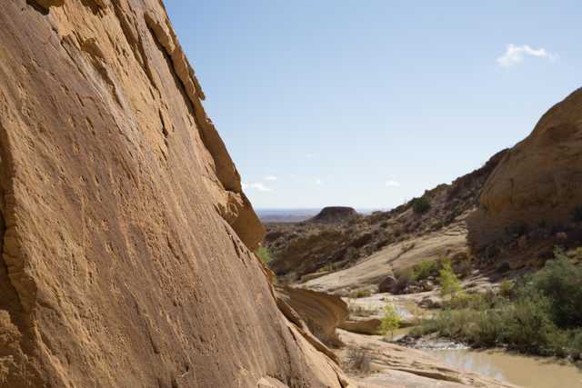 An arid landscape features large sandstone rock formations and a small stream in the foreground.